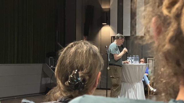 A speaker is at a podium, holding a microphone and addressing an audience. In the foreground, the back of a person's head is visible, along with another person seated in the background. The setting appears to be an indoor event space with a green curtain