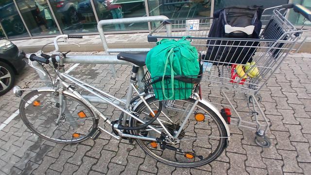 Ein altes silberfarbenes Fahrrad steht an einem Supermarkt-Radständer. Der Fahrradkorb ist mit Taschen gefüllt, dahinter steht ein Einkaufswagen  mit Rucksack und weiteren Lebensmitteln.