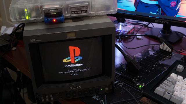A picture of a Sony PVM showing the SCEA PS1 logo on a black background with a crystal-shelled Playstation 1 on top.  Beside the PS1 is a gwscart switch with two SCART cables plugged in: one from the PS1 to the switch, and one from the switch into the PVM.