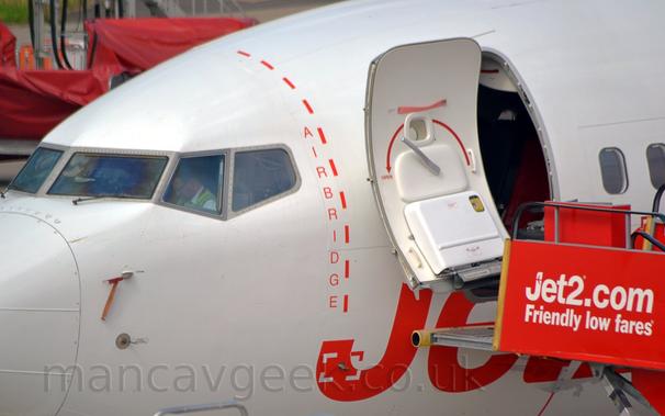 Close-up detail of a white jet airliner parked facing to the left showing the area around the open forward cabin door.
A red set of mobile airstairs with white "Jet2.com" above "Friendly Lower Fares" titles has been positioned by the open door, a yellow bumper leaning against the side of the fuselage just below the door.
Large, red "Jet2" titles are on the side of the plane just below the door, but are largely obscured by the airstairs.
Between the door and the cockpit windows, there is a dashed red line with the word "Airbridge" arranged vertically on the left.