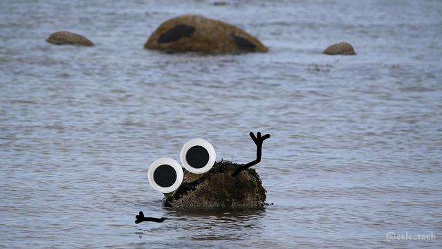 A photo of a calm sea with rocks poking out of it. The original photo had two. One in the foreground covered in barnacles and seaweed with a diagonal dark strip across it (like a mouth). One in the background, slightly out of focus, with two large black marks (like eyes). To the one at the back I added two smaller rocks, like hands starting to reach out. To the one in front I added large googly eyes, and upturned arms. I'm not sure what's really going on, but the one with the big black marks did really look like some kind of stone golem peering out of the sea.