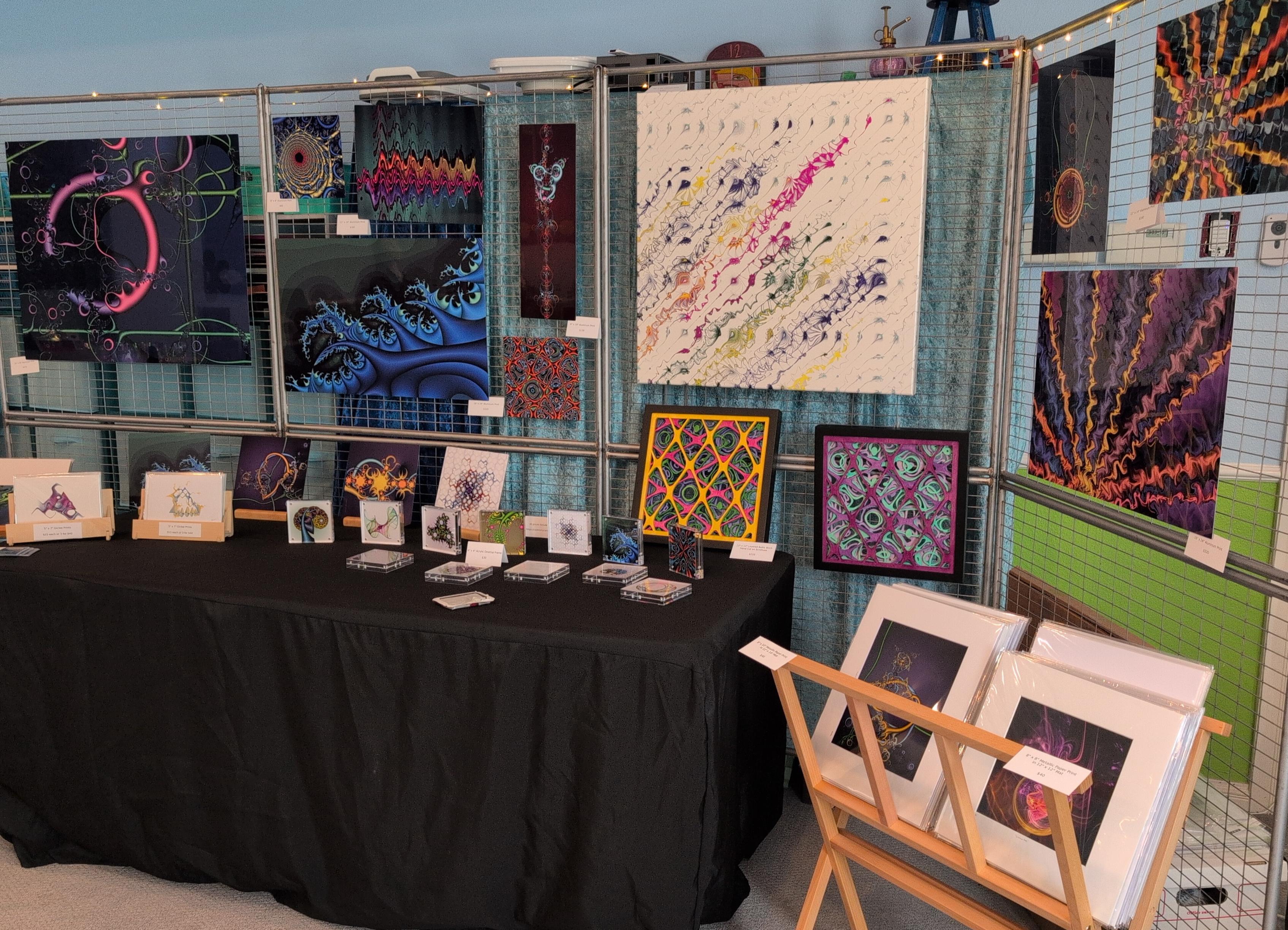 A display of fractal and algorithmic artwork with pieces of various sizes and colors hanging on the wall and on a table, ready for a show.