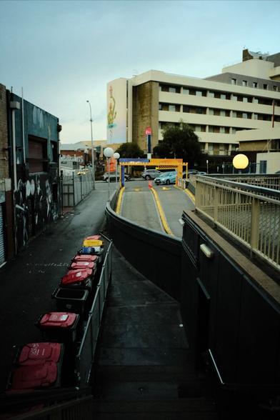 A cityscape on a rainy day. Rubbish bins with red and yellow tops are visible in the lower left. A yellow gate to a parking garage appears in the the middle of the frame. A large mural appears on a building in the background with a human like figure. The sky is cloudy and hinting at rain.