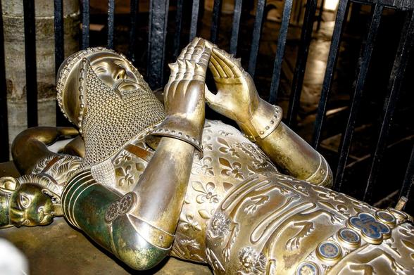 A metallic sculpture of a medieval knight in full armor reclines with interlaced hands near his chest and a peaceful expression on his face. The knight wears a detailed helmet with chainmail, ornately engraved armor showcasing floral patterns, and jointed gauntlets. His legs are slightly bent, and his feet rest near a three-dimensional lion head, part of the sculpture. The setting includes a dark background with metallic bars, possibly indicating an indoor location.
