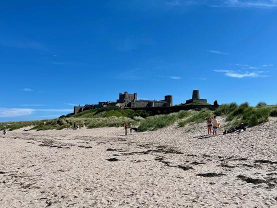 Golden sand stretches across Bamburgh’s coastline, framed by gentle dunes topped with tufts of green grass, while the imposing silhouette of Bamburgh Castle rises against a brilliant blue sky. Along St Oswald's Way, beachgoers stroll, sunbathe, and enjoy the wide, open expanse, with a few people and dogs exploring the shoreline. The castle’s stone walls and towers dominate the horizon, their weathered structure contrasting with the vivid colors of the landscape, evoking centuries of history overlooking the North Sea. Wisps of seaweed are scattered across the sand, and the light breeze carries a sense of calm as the vast sky stretches uninterrupted above.