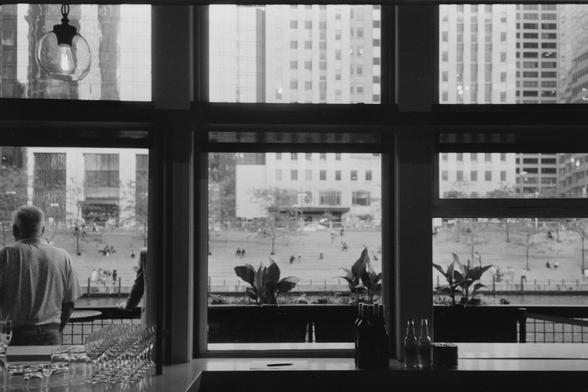 Black and white photo taken from indoors looking through a set of windows each panel taking a third of the frame. The interior has counters with glassware on the left and glass bottles in the middle. The exterior has some potted plants,a person standing with their back to the camera, and in the distance across the river a bank of steps can be seen with specs of people scattered about.