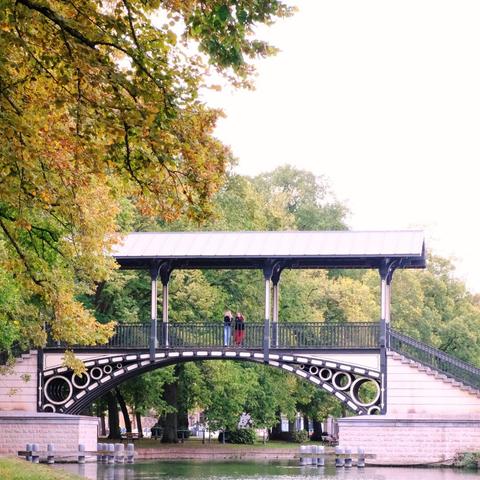 Covered pedestrian bridge with decorative arches crossing over a calm canal, surrounded by green and golden trees. Two people stand at the center of the bridge.