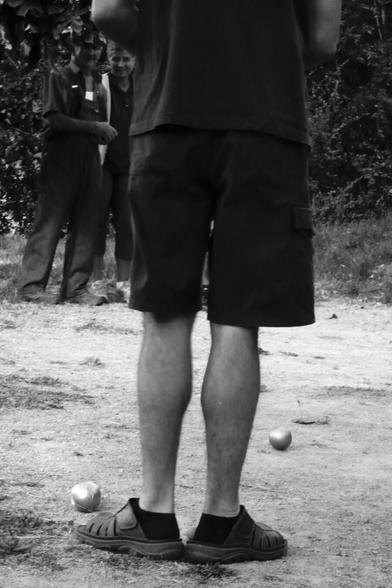 One player stands with er feet behind the jack on the pétanque strip, indicating the target to his teammates.  (Version françæ) Æn personne se tient les pieds en canard derrière læ cochonnet sur æn terrain de pétanque pour indiquer læ cible à ses coéquipiærs.
