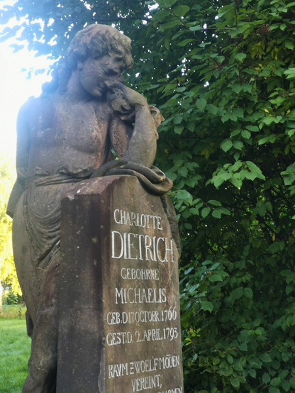Nahaufnahme eines alten Grabsteins mit einer steinernen Frauenfigur. Die Figur sitzt mit geneigtem Kopf und legt eine Hand an ihr Gesicht, als würde sie trauern. Auf dem Grabstein steht in weißen Buchstaben: ‚Charlotte Dietrich, geborene Michaelis, geboren am 17. Oktober 1766, gestorben am 2. April 1793‘. Der Grabstein ist von dichtem grünen Laub umgeben.