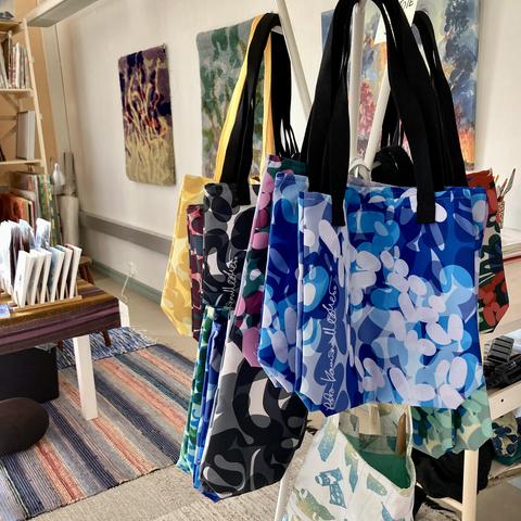 An interior shot of an art studio with a coat rack of colourful art print tote bags in the foreground, a table with art products, and woven wall hangings and a painting hanging on a wall.