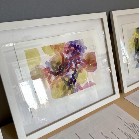 A close-up of an abstract microscopy watercolour that is framed behind glass in a white frame and leaning against a grey wall. Sunlight from a tall window is reflected in the glass.