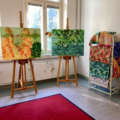 An interior shot of a corner in a wide corridor in which are situated two wooden easels with medium-sized abstracted landscapes in blue, yellow, and green. Next to the paintings on the right is a wooden frame with folded meters of different colourful printed fabric.