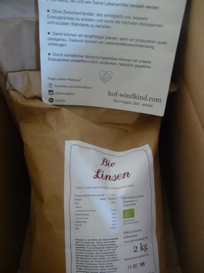 Der Blick in einen etwas dunklen Karton. Eine Papiertüte mit Bio-Linsen. Dahinter ist noch eine zu erahnen. Oben liegt, halb zu sehen, ein Zettel von Hof Windkind mit einem Teil ihrer Aufzählungen, für was die Feldadoptionen gut sind. Ohne Zwischenhändler, Planbarkeit und einheitliche Verpackungsgrößen. Dann steht da noch, wo sie online zu finden sind. Leider noch nicht im Fediverse.