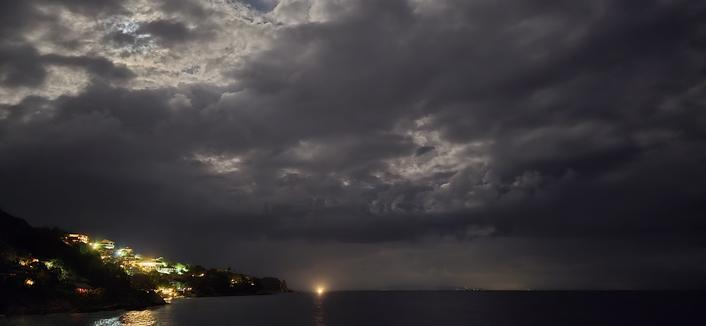 photo de nuit de la mer en bas, des collines qui descendent s'y jeter en bas à gauche et de quelques lumières au large en bas à droite. Les deux tiers supérieurs de l'image sont occupés par des nuages sombres, au travers desquels la lumière de la lune peine à passer