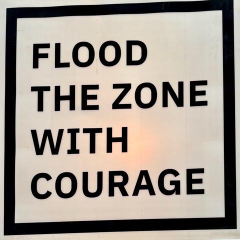Ein weißes Schild mit schwarzem Rahmen. In großen schwarzen Buchstaben steht: „FLOOD THE ZONE WITH COURAGE“.