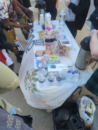This is a photo of the Wednesday Morning coffee serve. A view from the side of the serving table, with people to the left (front of the table) crowded around getting food and people to the right (behind the table) crowded around serving. Spread across the table are coffee-serving pots, water bottles, masks, hygenic products, PBJs, energy bars, and the coffee supplies like oat milk and cups.