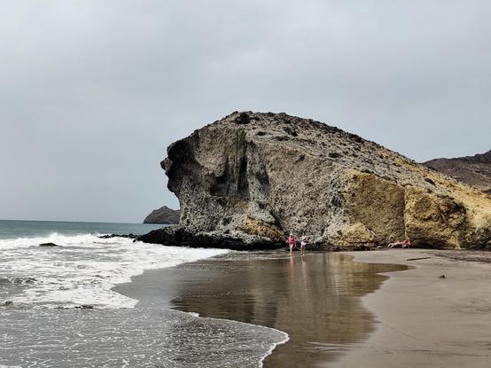 El morrón de Monsul desde la playa. Las olas rompen contra las rocas y algunas personas pasean por la playa.