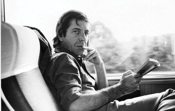 A black and white photograph depicts Leonard Cohen sitting in a train seat, holding a book. He is looking directly at the camera with a contemplative expression. Courtesy Old Ideas, LLC