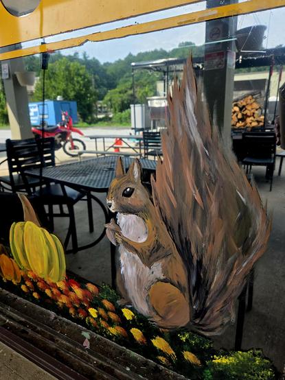 This is a painted mural on a glass window, showing a lively fall scene. The main subject is a large squirrel with a bushy tail, sitting upright as if nibbling on something with its tiny front paws. The squirrel is painted in warm browns, tans, and whites, with textured brushstrokes giving life to its fur. Beside it are bright autumn pumpkins in yellow and orange shades. In front, a patch of colorful flowers—red, orange, and yellow—adds a vibrant base. Through the glass, the outdoor setting is visible: black patio chairs and tables, a stack of firewood, and a red motorcycle parked near a building, all under a sunny sky with green trees in the background. The painting feels festive and seasonal, celebrating autumn.