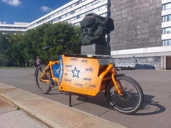 Ein oranges Lastenrads steht vor einem riesigen Kopf aus Bronze, das Karl-Marx-Denkmal.