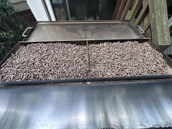 A close-up view of a large, open metal hopper filled with wood pellets. The hopper is divided into two sections, each brimming with cylindrical brown pellets used as fuel in a pellet grill or smoker. The surrounding surface of the grill is metallic with visible discoloration from use, and part of a wooden deck and brick wall are visible in the background.