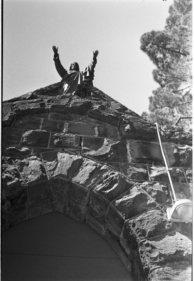 A black and white view of an abandoned church entrance above which a Jesus figure has outstretched arms as though about to dive.