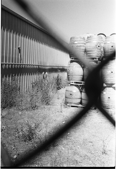A stack of barrels viewed through a blurred chainlink fence link.  Black and white.