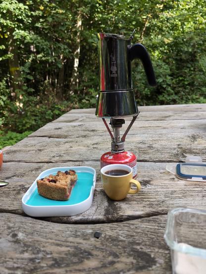 Eine Bialletti auf einem kleinen Campingkocher, eine gelbe Espressotasse und ein Stück Zwetschgenkuchen auf einem Holztisch im Wald.