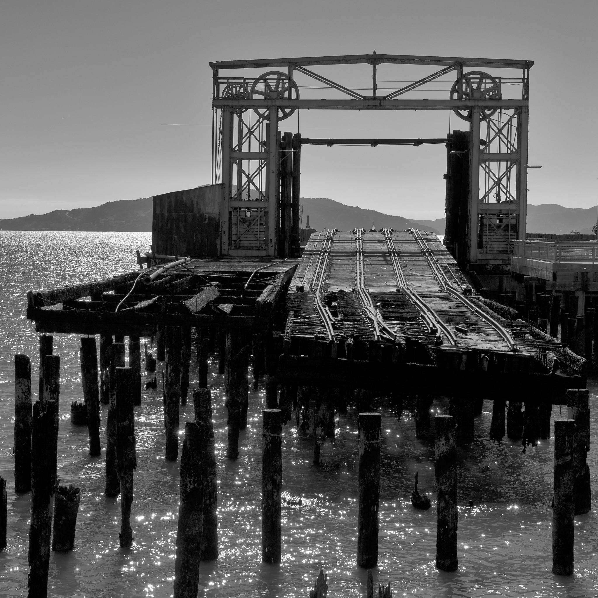 Charred ties and tracks on a pier over water, with hills across the water in the background.