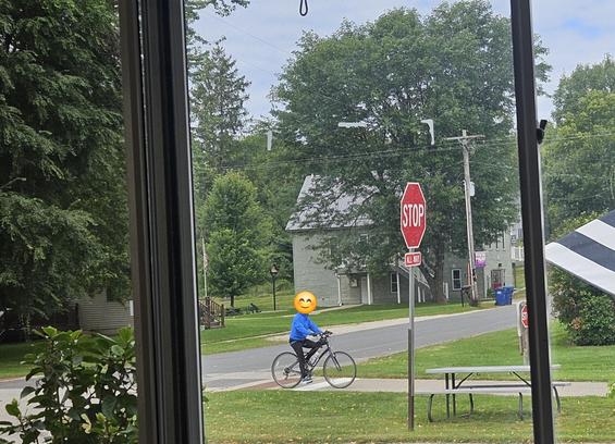 A bicyclist on an asphalt trail, crossing an asphalt street, green grass on both sides. A red stop sign is visible. A wood park bench is on the side of the trail. A 2 story light green house is in the background.