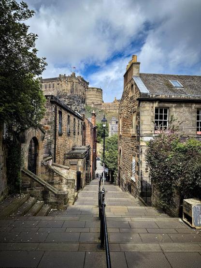 Narrow cobblestone street lined with historic stone buildings leading to a large castle on a hill under a cloudy sky. Green foliage on the left, metal railing along the descending path, and scattered leaves on the ground.