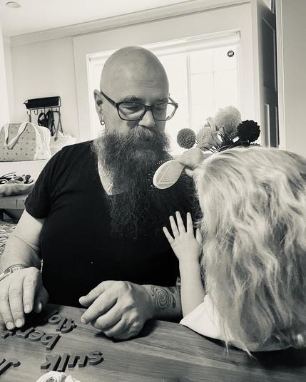 A black and white photograph of a young girl brushing her bald grandfather’s long beard.