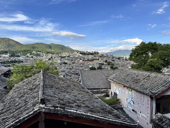 Old-style roofs, off into the distance in a Chinese mountain town. A blue sky with a few clouds, and a mountaintop in the distance to the left. In the wall of a building towards the right & front of the frame is some painted writing and pictures.