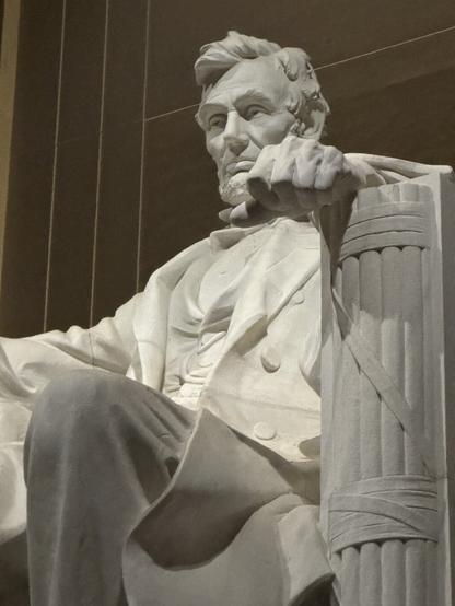 A large marble statue of a seated Abraham Lincoln. The figure has a thoughtful expression, with one hand resting on a column. The background features dark architectural elements, and the overall tone is solemn and grand.