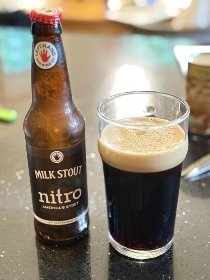 A brown bottle with a black label and white lettering is topped with a red bottle cap. A pint glass of dark beer has a creamy light tan head.