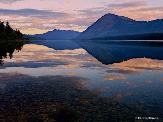 A calm sunrise scene at Lake Wenatchee, Washington in 2023. The glassy surface of the lake creates a perfect mirror reflection of the mountain ridge, pink and gold clouds, and surrounding tree line. The foreground reveals the rocky lake bottom beneath the clear water, while the horizon glows with soft pastel hues of dawn. The stillness of the water makes the sky and mountain appear seamlessly doubled, producing a striking symmetrical composition.