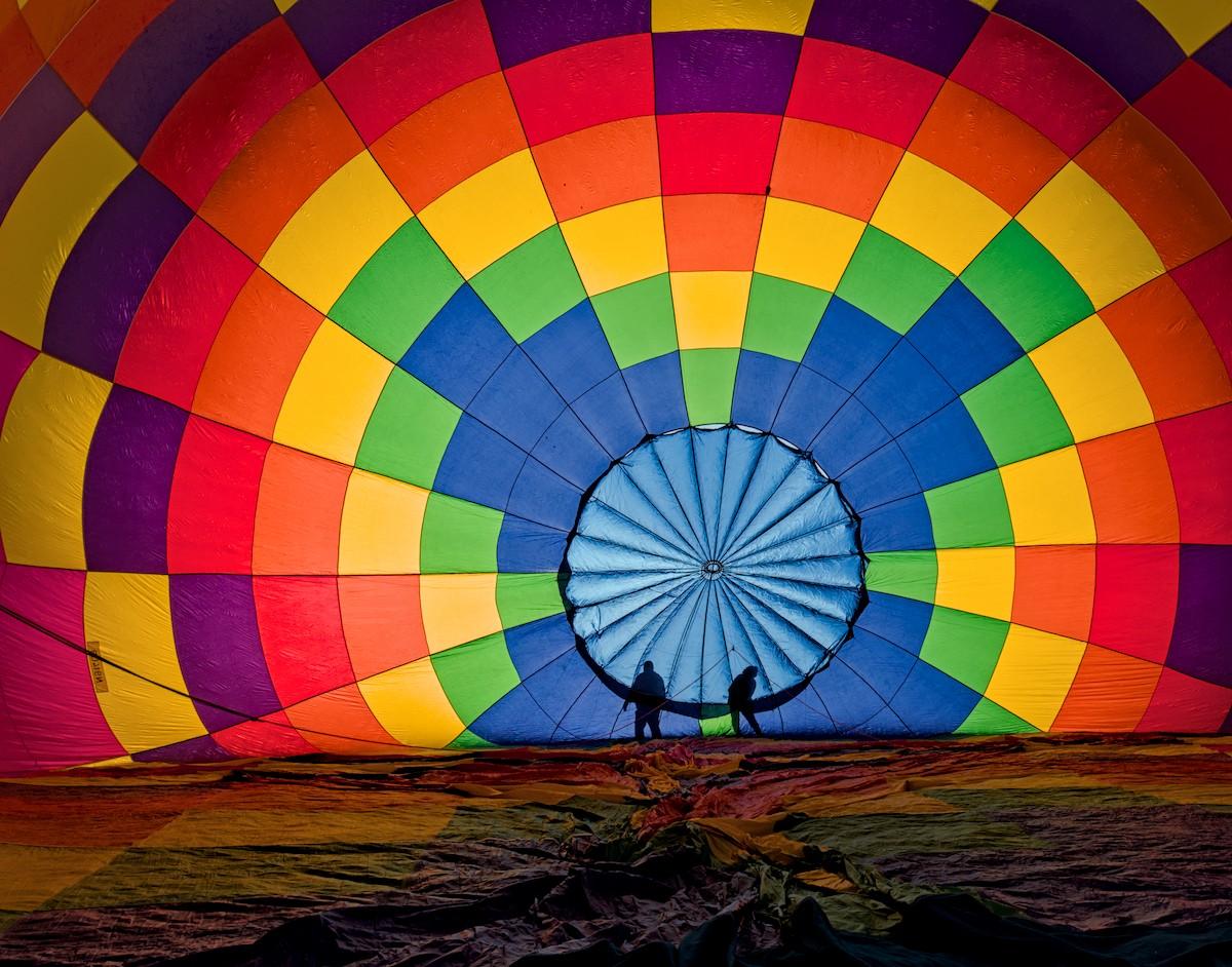A color landscape photo of the view looking into the envelope of a hot air balloon as it is beginning to inflate. It is a checker pattern of blue, green, red, orange, pink, and purple. The round top vent is light blue with the early morning sun lighting it from behind. The silhouettes of two people, small against the large balloon are seen on the ground adjusting some of the fabric as it inflates.