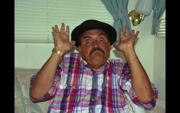 Gaspar Henaine, conocido como Capulina,haciendo una expresión cómica, con una camisa a cuadros colorida y un sombrero negro, en un ambiente interior con ventanas y cortinas.