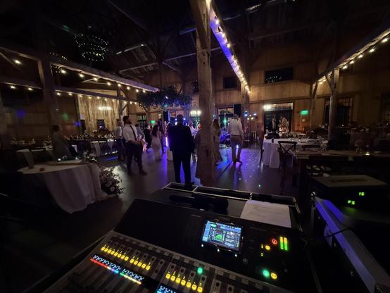 Image looking over a digital mixer setup in a barn with hanging lights. A live band plays for wedding guests.