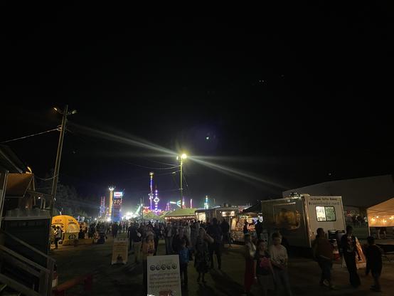 Dark nighttime shot of the fair shows people streaming towards the camera down a wide path between buildings and food, stands with a midway and bright lights in the background