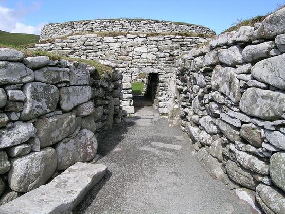 Clickimin Broch. The image shows a gravel path leading between high walls made of large very light grey stones. It leads to a rectangular doorway in a large circular structure, also made of unmortared stones. There is a thin layer of turf on top of all the walls. The sky is blue.