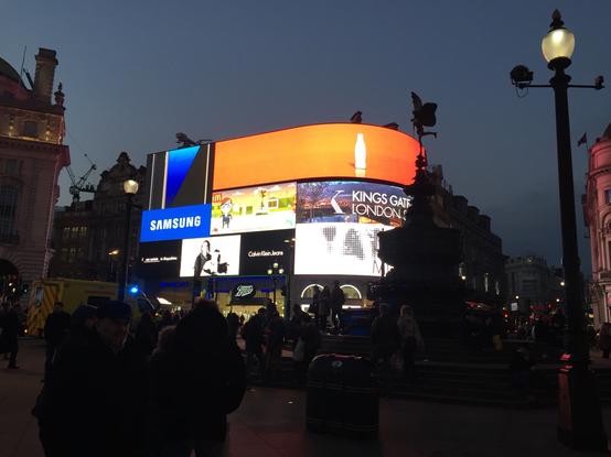 Eine geschäftige Stadtszene in der Abenddämmerung mit beleuchteten Werbebildschirmen am Piccadilly Circus in London. Prominente Marken wie Samsung und Coca-Cola werden angezeigt. Eine Menschenmenge versammelt sich in der Nähe eines Brunnens, mit einigen Rettungsfahrzeugen im Hintergrund.