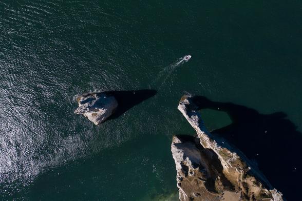 Aerial view of rocky formations jutting out of a dark green sea, with a small boat leaving a wake as it moves through the water.
