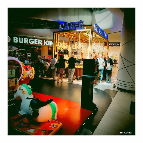 Schnappschuss aus einem Einkaufszentrum mit Leuchtreklamen für ein Café-Kiosk sowie bekannte globale Unternehmen in Fast Food und Unterwäsche. Das Café ist von Kunden umringt. Im Vordergrund links ein kleiner Spielplatz für Kinder. Vignette for Android auf Xiaomi Redmi 14+.
