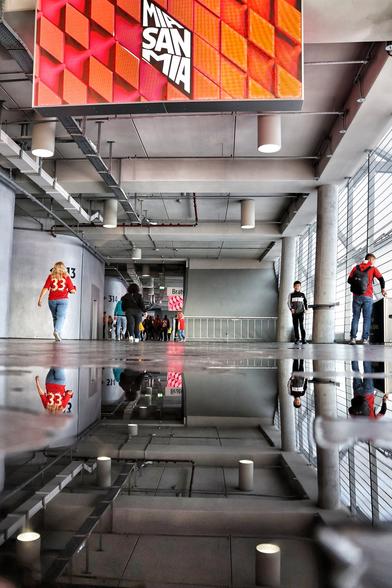 Das Bild zeigt das Innere eines Stadions (die Allianz Arena). Der Blick fällt auf einen breiten, offenen Gang mit einer Pfütze auf dem Boden, die die Umgebung und die Personen darauf deutlich reflektiert. Die Architektur ist industriell geprägt: Die Decke ist offen gestaltet und zeigt sichtbare Rohrleitungen sowie Beleuchtungselemente. Massive Betonpfeiler gliedern den Raum. An der Decke hängt ein großes rechteckiges Schild mit einem geometrischen Muster in Rot- und Orangetönen. Darauf steht in großen weißen Buchstaben „MIA SAN MIA“. Entlang der Seitenwände sind nummerierte Bereiche wie „313“ und „314“ zu erkennen, die vermutlich auf Sitzplatzbereiche hinweisen. Mehrere Personen bewegen sich durch den Gang, einige davon haben ein rotes Trikot an.