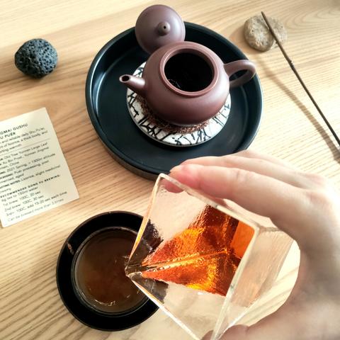A gong fu tea set seen from above. In the background is a yixing small open teapot on a ceramic water tray, and next to it are a small black stone, a paper with some information about the tea, and a small branch used to push the tea in the teapot. In the forefront is a dark, small tea cup, and my hand holding a pitcher and pouring the tea from it into the cup. The tea is bright orange.