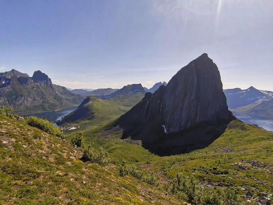 The image depicts a stunning mountainous landscape under a clear blue sky. In the foreground, there is a grassy slope with patches of green vegetation and small yellow flowers. The middle ground features a prominent, steep, and rugged mountain peak with a sharp, pointed summit, which is likely the main focus of the image. This mountain has a dark, rocky surface with a small waterfall cascading down its side. To the left, a river or fjord winds through the valley, bordered by more green hills and mountains. In the background, additional mountain peaks are visible, some with snow caps, indicating higher elevations. The sunlight casts shadows on the mountains, enhancing the texture and depth of the scene. The overall composition highlights the natural beauty and grandeur of the landscape.