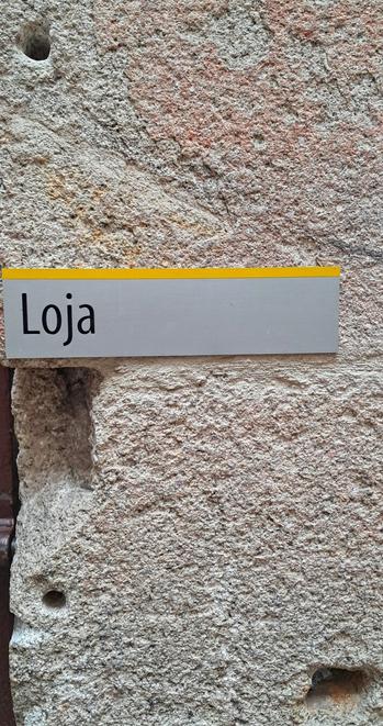 [pt] Parede de pedra irregular e com buracos. Ao meio uma placa de metal a dizer Loja

[en] Irregular stone wall with holes. In the middle is a metal plaque saying Loja