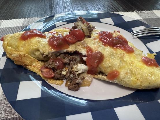 A golden-brown omelet folded over a hearty filling of ground beef, onions, and sliced pepperoni, topped with melted cheese and spoonfuls of chunky tomato sauce. The omelet sits on a blue and white checkered plate, with some of the savory filling spilling out from the inside. A fork rests to the side on the plate, ready for eating.