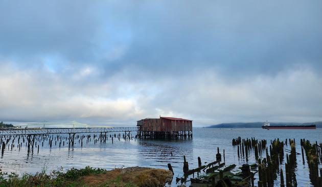 A color photo of the waterfront in Astoria, Oregon. In the foreground at the bottom are old pilings and a small spit of land. In the river is an old building with a red roof and a narrow rickety pier leading to it. In the background left is a big bridge. In the background right is a huge ship. The sun is coming through the clouds and illuminating the big bridge.